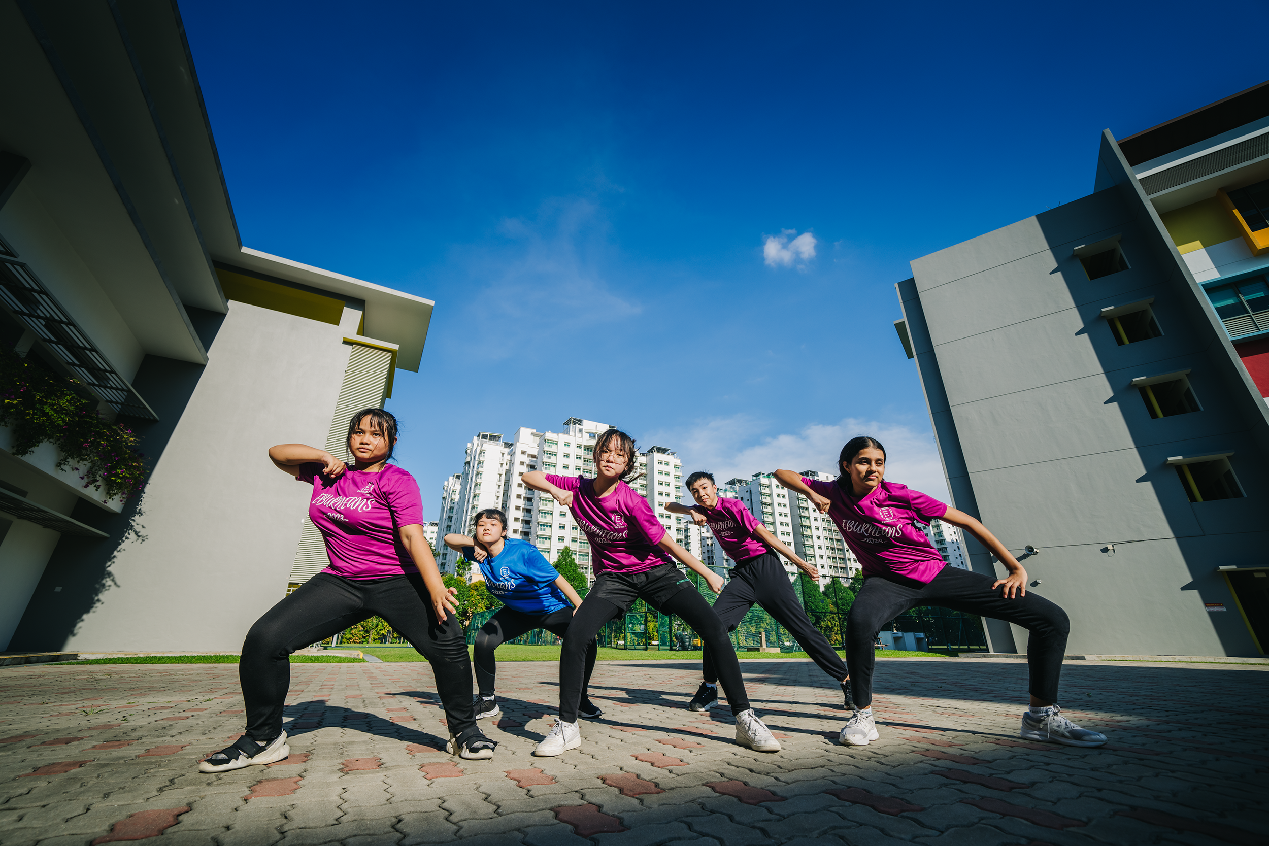 Five dancers in pink and blue shirts pose in a courtyard between buildings under a bright blue sky.